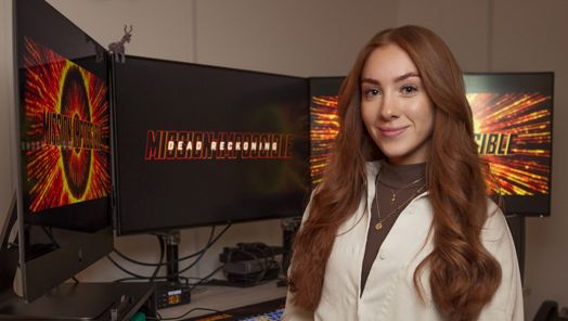 A woman stands smiling towards the camera in front of three monitors displaying the Mission Impossible logo in large bright colours.
