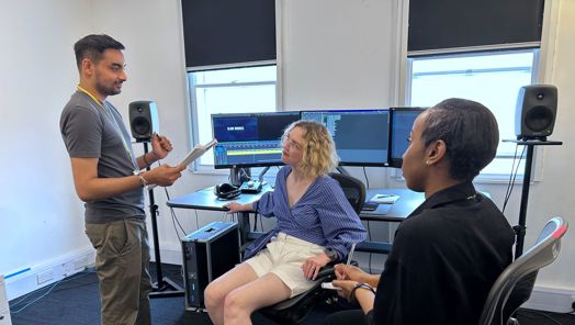 A person, stood, gives instructions to two other people, seated, in a room set up behind them with a set of computer monitors upon the desk and speakers on either size.