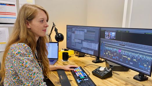 A woman sits at a desk in front of a set of computer monitors editing a sequence in a video editing software.