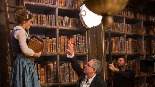 A scene in a library where a director instructs an actress holding some books. In the background, a lights operator adjusts the lighting.