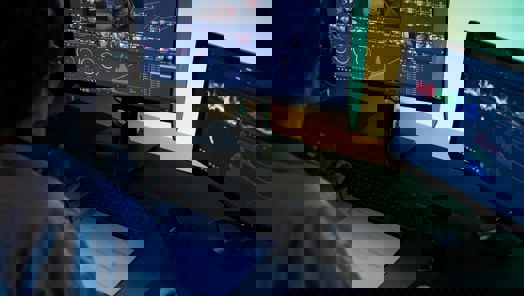 A person sits at a desk in front of two monitors editing the colours of a video onscreen 