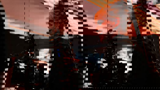 A woman holds a large camera which is strapped to her on a film set