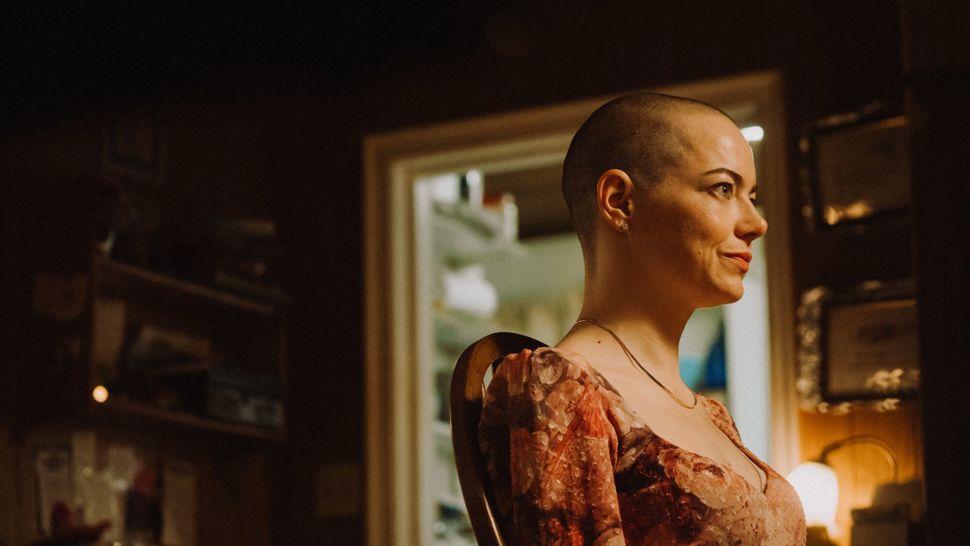 Emma Stone stands in a dimly light room looking out towards the right.