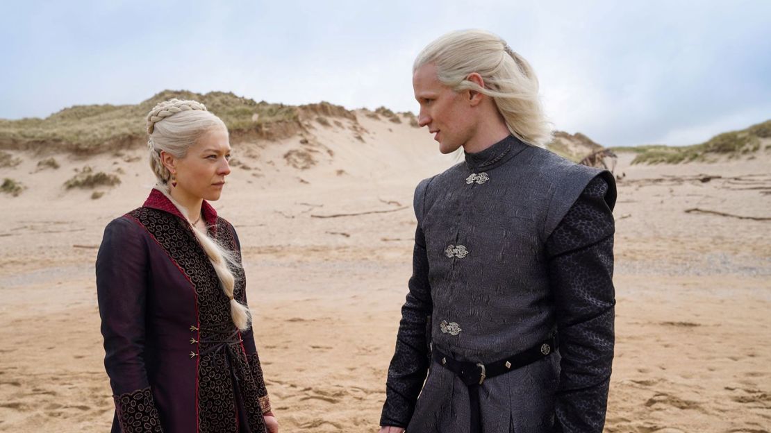 Two characters from HBO series House of the Dragon stand pensively looking at each other on a beach with sand dunes behind