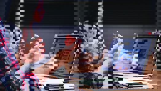 Close up of a person sitting at a large wooden desk in front of a laptop with a notebook and mobile phone on the desk