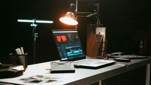 A laptop is open on a desk surrounded by books, papers and equipment. An overhead lamp shines on the screen which is open to a video editing software.