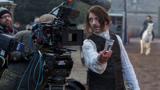 On the set of period drama, a man stands holding a glass. He looks towards a large film camera.