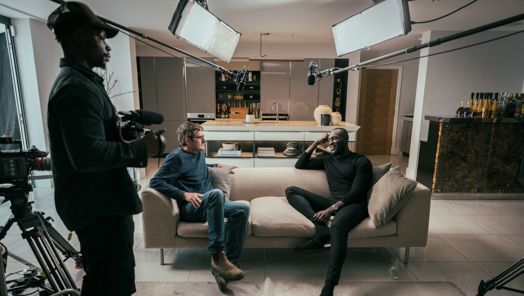Louis Theroux and Stormzy sit chatting on a sofa surrounded by filming equipment. A cameraman holding a camera stands off to one side.