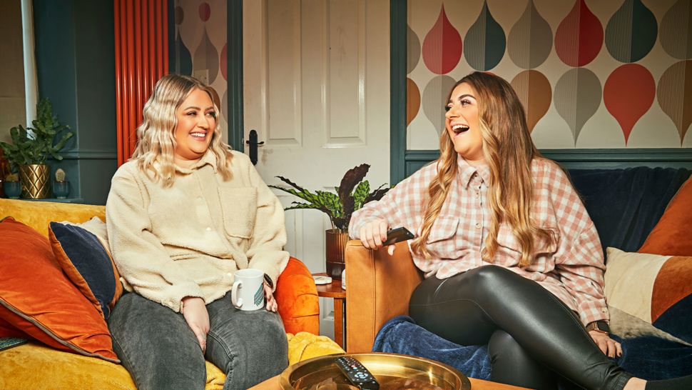 Production image of two Gogglebox viewers sitting on sofas smiling at each other with a TV remote on a tray in front of them