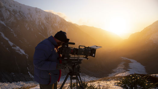 Camera operator is shooting a scene in the snow, wearing a heavy jacket and a hat.