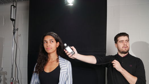 Young man holds out lighting device next to head of young woman on a set