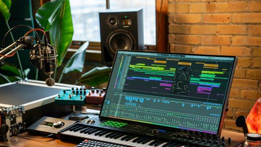 A screen monitor is set up on a desk, open to a sound editing software. Around the monitor is further sound equipment including a microphone in front of it.